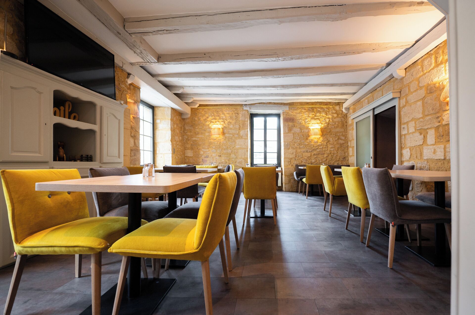 Een restaurant met stenen muren, houten balken en gele stoelen. Tafels staan in rijen, met een centraal geplaatst raam.