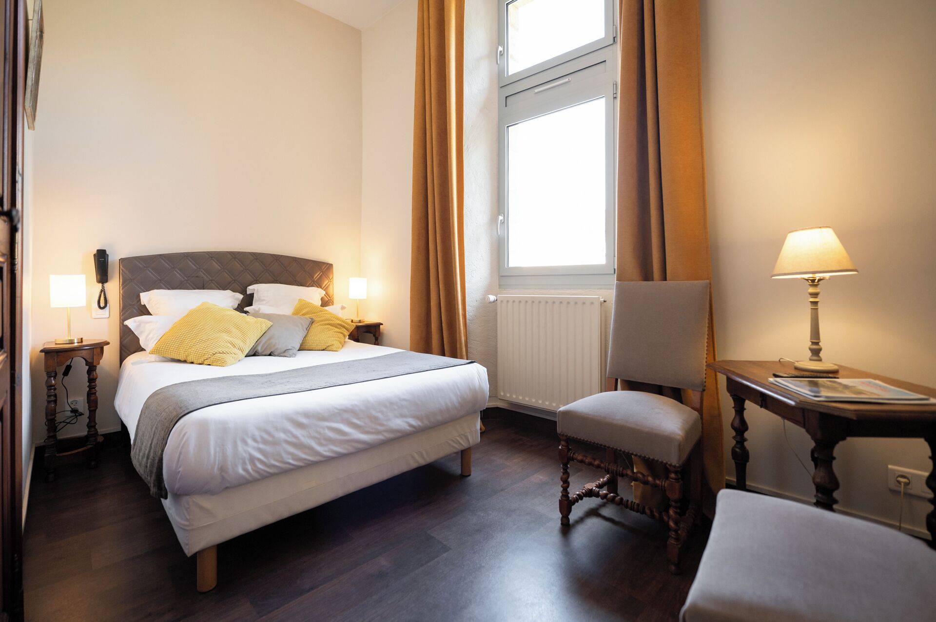 Hotellihuone, jossa on sänky, yöpöydällä lamput, tuoli, kirjoituspöytä lampun kanssa, ikkuna verhoilla ja seinässä puhelin.