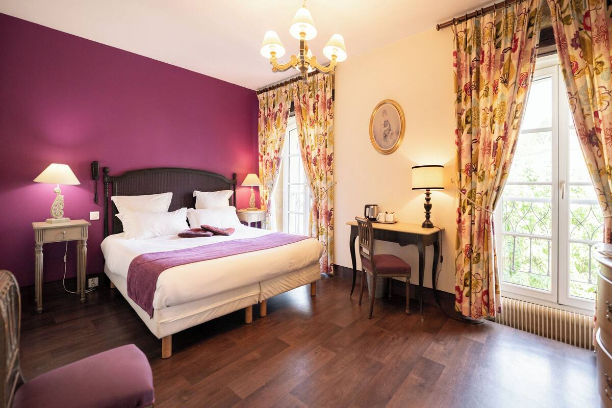 A hotel room with a bed, bedside tables, a desk, a chair, and a window with curtains.