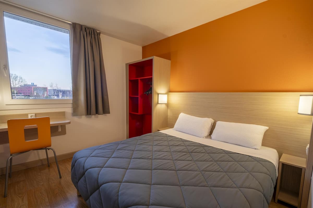 A hotel room with a large bed, a desk with a chair, a red storage unit, and a window with curtains.