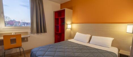 Una habitación de hotel con una cama grande, un escritorio con silla, un mueble de almacenamiento rojo y una ventana con cortinas.