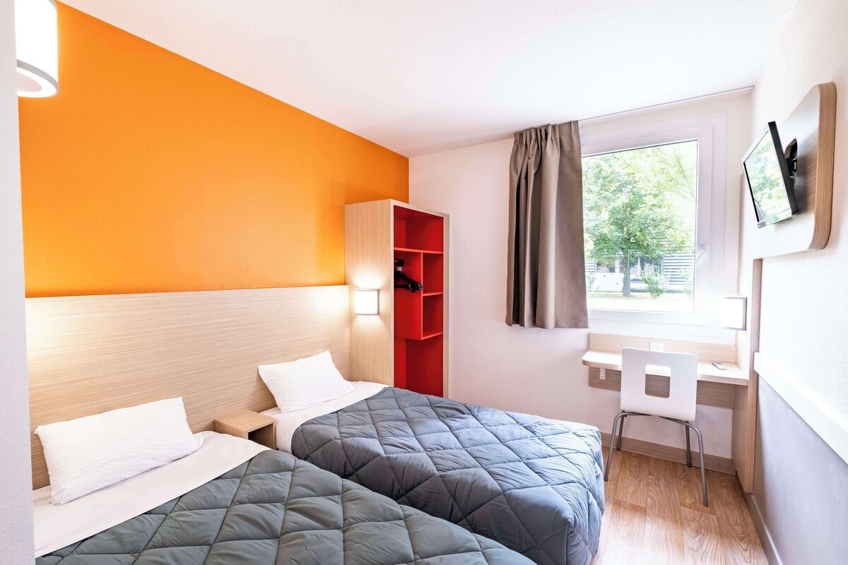 A hotel room with two beds, a desk, a chair, a TV, and a window with curtains.