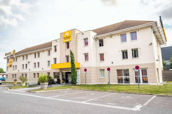 A hotel with a yellow sign indicating a premiere class. It has multiple floors, a parking area, and a small garden with potted plants. - Premiere Classe Roissy - Aéroport CDG - Le Mesnil Amelot (Le Mesnil-Amelot)