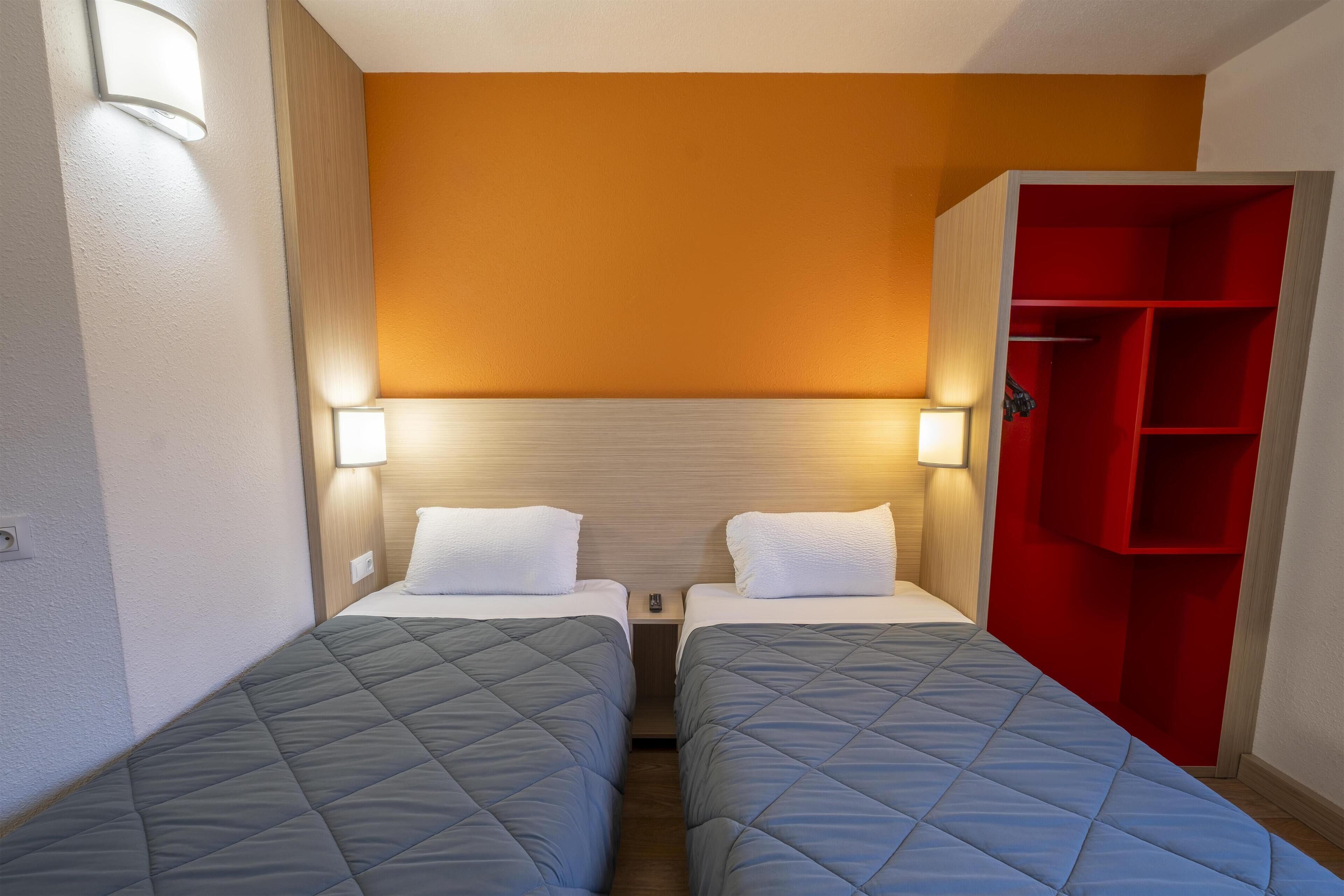 A hotel room with two beds, a red wardrobe, and a wall-mounted light fixture.