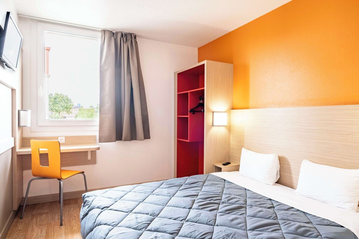 A hotel room with a bed, a desk, a chair, a wardrobe, and a window with curtains.