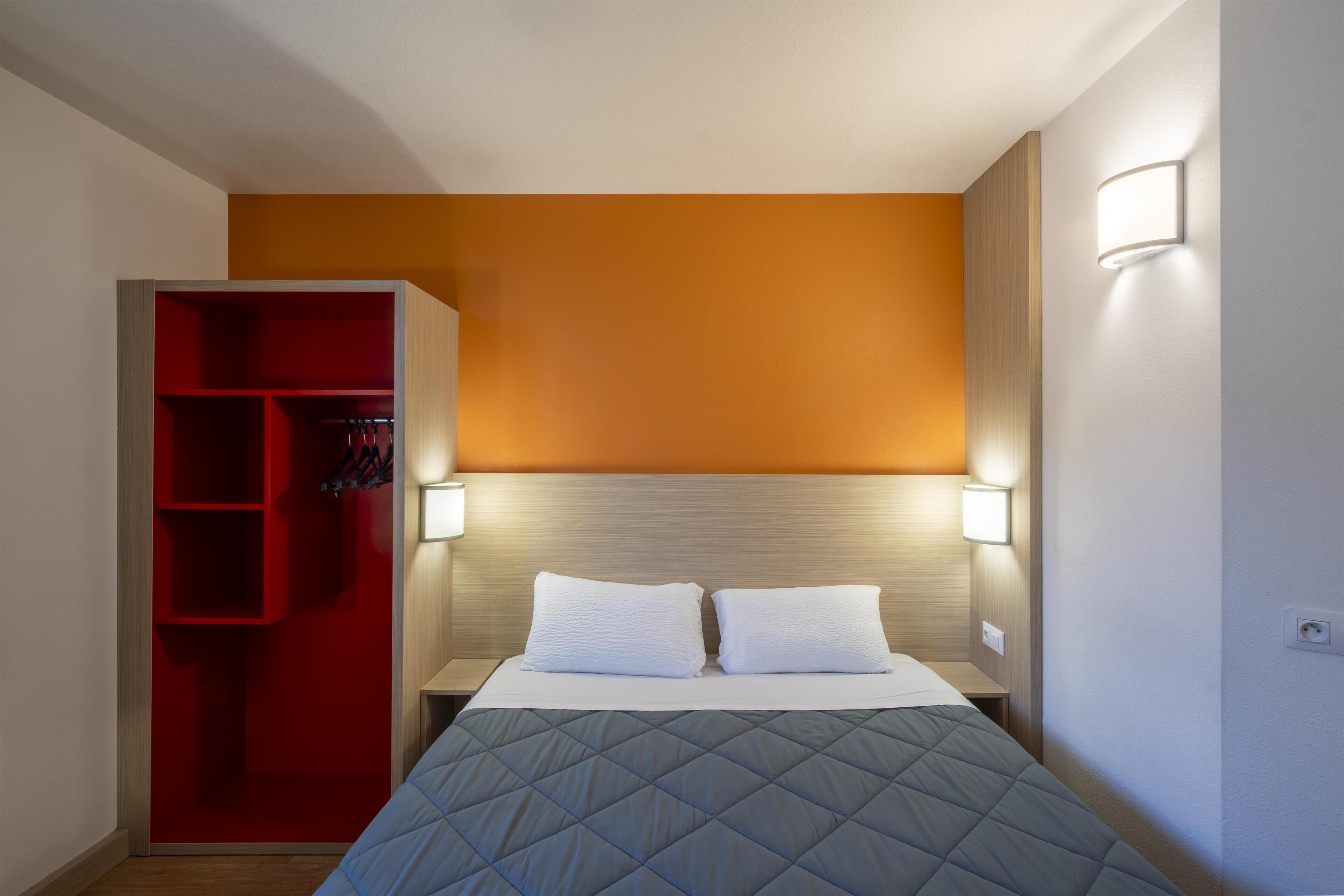 A hotel room with a bed, two bedside lamps, a red wardrobe, and a wall-mounted light fixture.