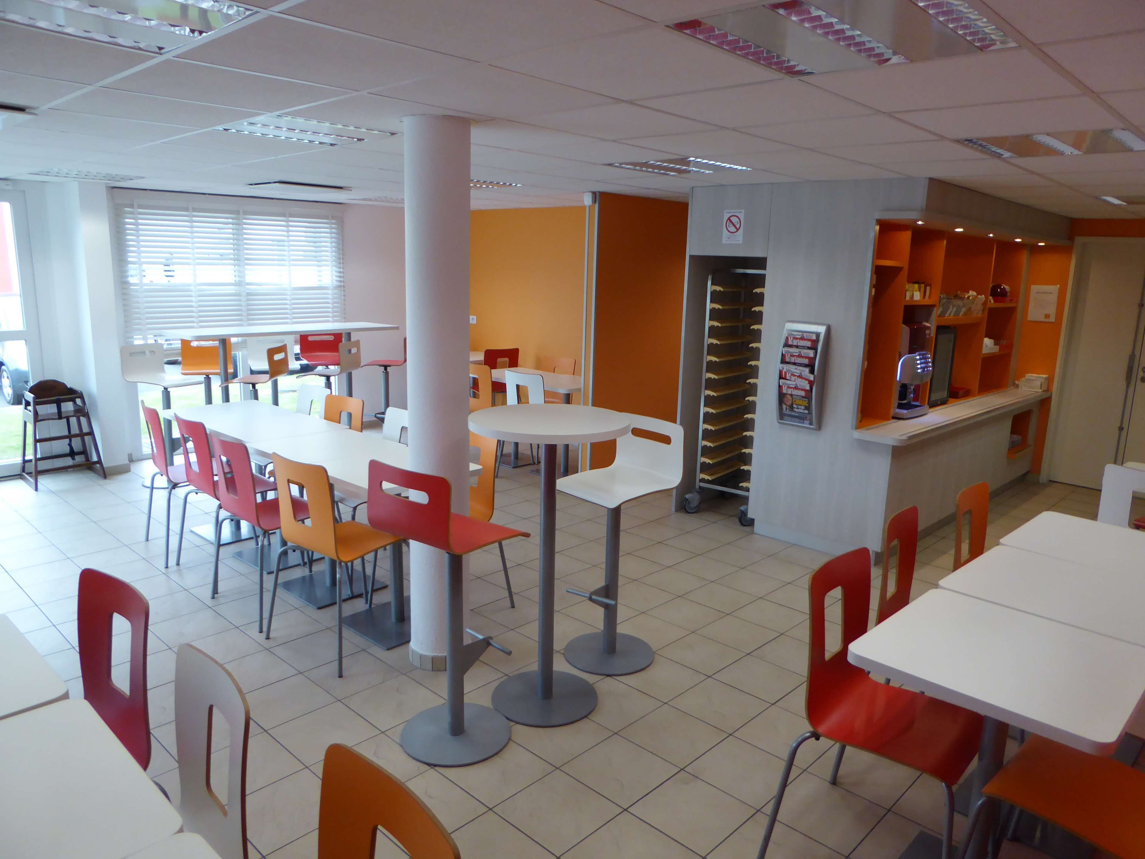 Eine Cafeteria mit weiß-orangenen Tischen und Stühlen, ein Getränkeautomat und ein Rauchverbot-Schild.