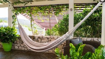 Una hamaca bajo una pérgola de madera con paredes de piedra, muebles de mimbre y plantas en macetas.