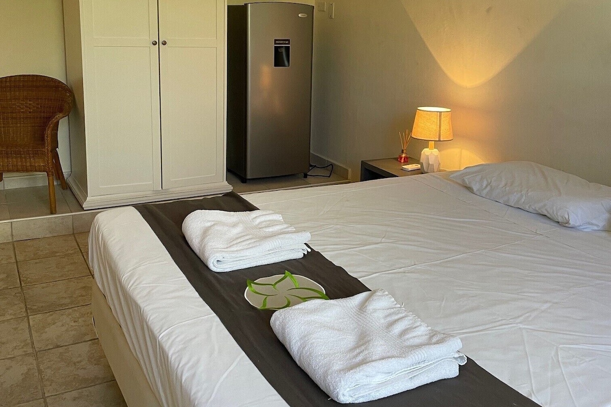 A hotel room with a bed, a wardrobe, a refrigerator, a chair, and a nightstand with a lamp.