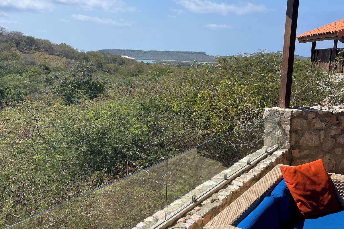 A balcony with wicker furniture and a stone wall, overlooking a hilly landscape with a clear sky.