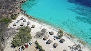 Vue aérienne d’une plage avec des huttes aux toits de chaume, une eau turquoise cristalline et un espace de stationnement.