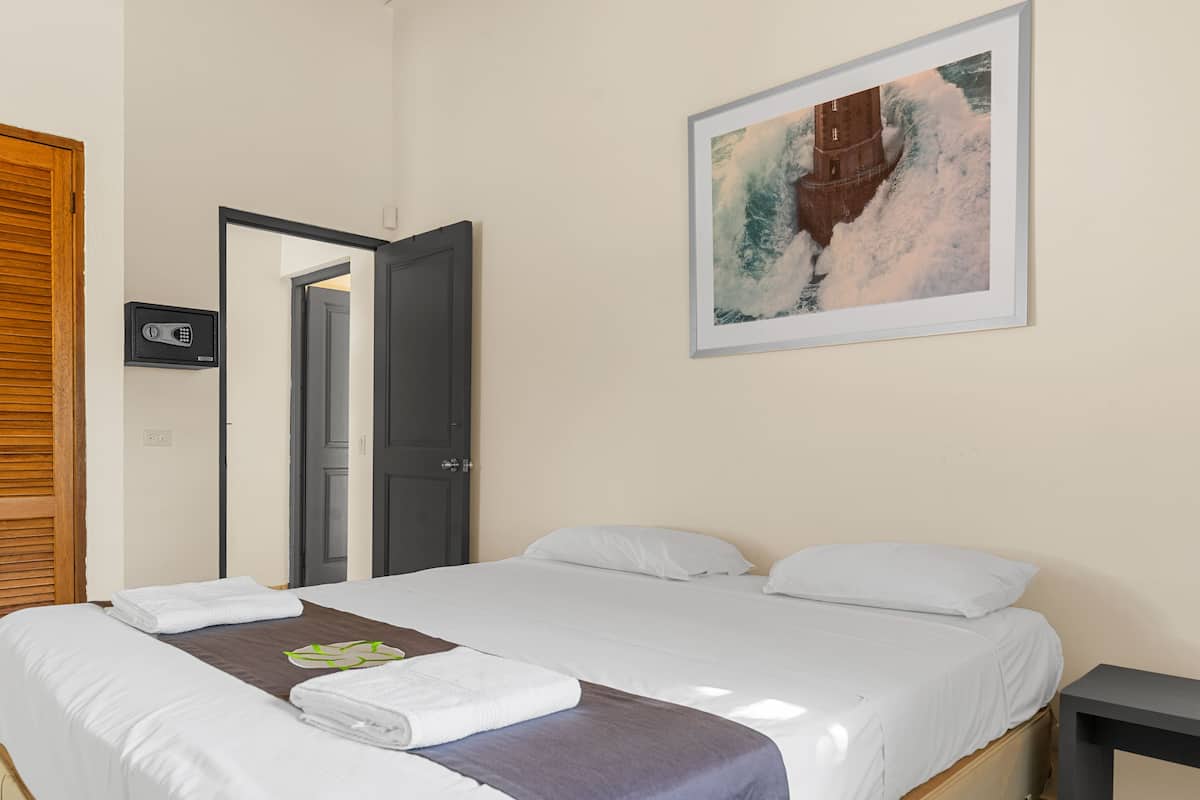 A hotel room with a bed, a painting of a lighthouse on the wall, a door, and a small table.