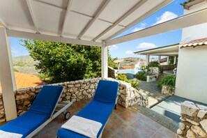 A covered outdoor area with two blue lounge chairs, a stone wall, and a view of a pool and hillside. - Bandabou Breeze Curacao (Sint Willibrordus)