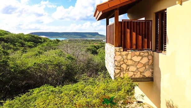 Une maison avec des fondations en pierre et un balcon en bois donnant sur un paysage verdoyant et offrant une vue lointaine sur une étendue d’eau.