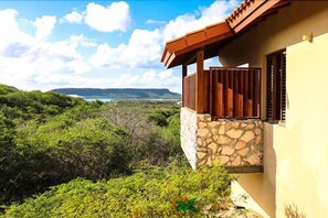 A house with a stone foundation and a wooden balcony overlooking a lush green landscape and a distant view of a body of water. - Bandabou Breeze Curacao (Sint Willibrordus)