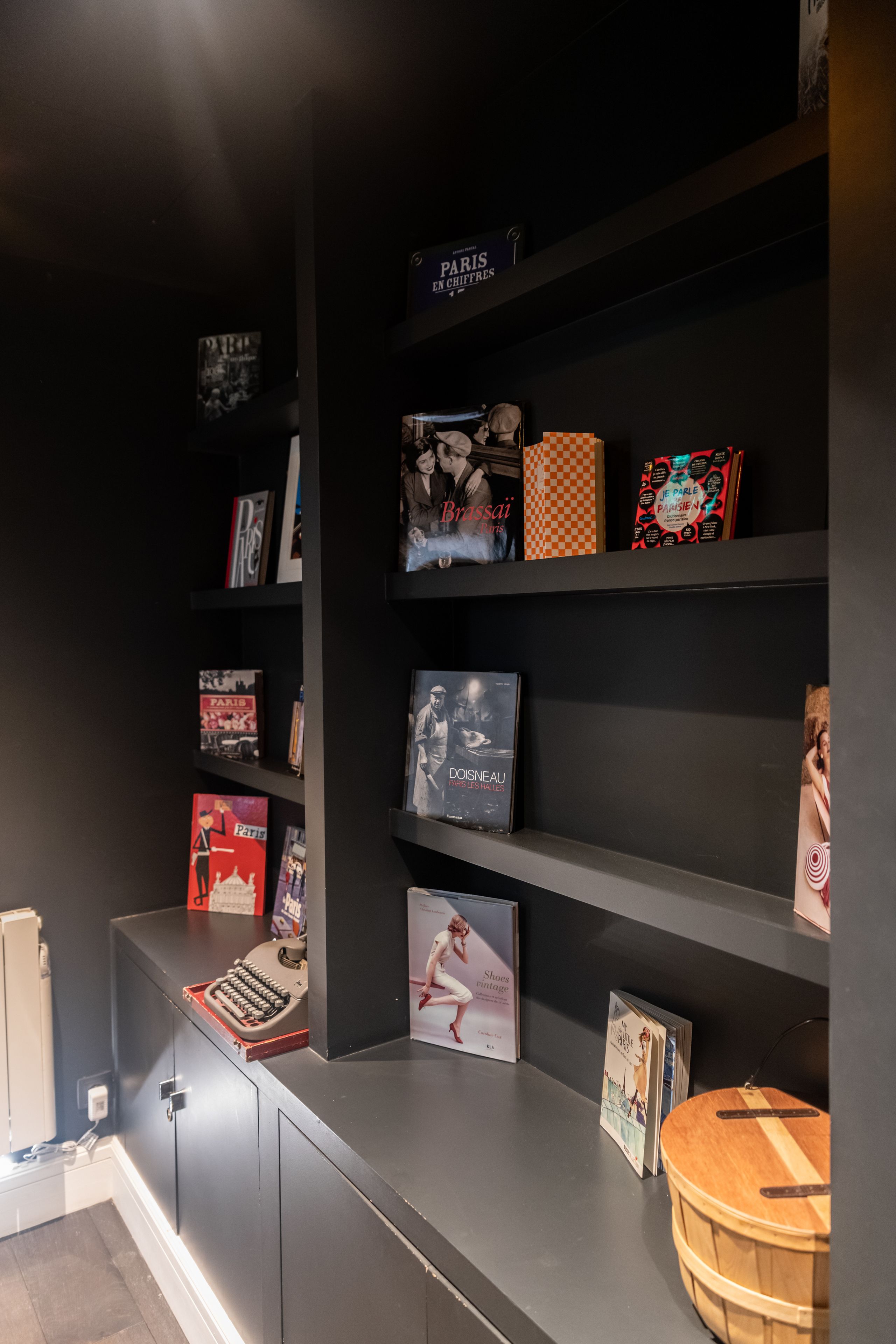 A dark-themed room with a bookshelf containing books and magazines, a typewriter, and a wooden bucket.