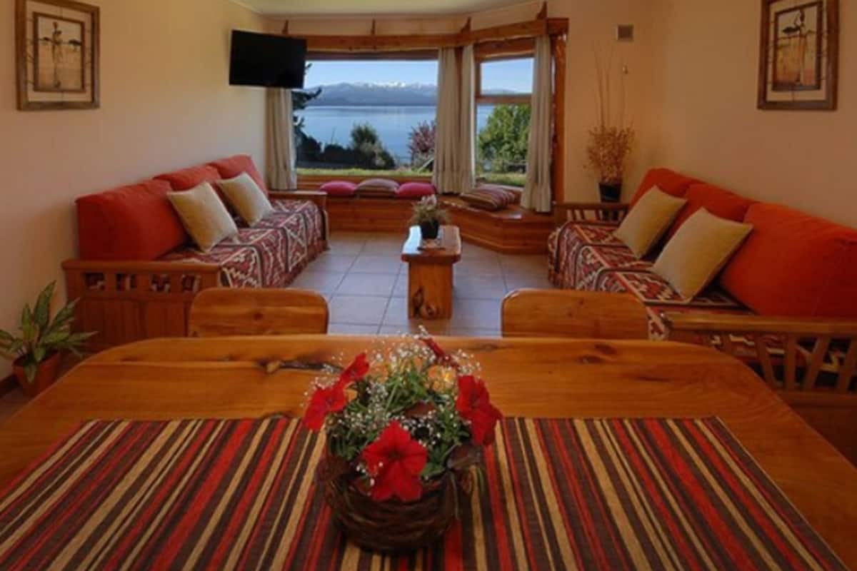 Un salon chaleureux avec une grande table en bois, deux canapés ornés de coussins rouges et une vue sur un lac, visible à travers de grandes fenêtres.