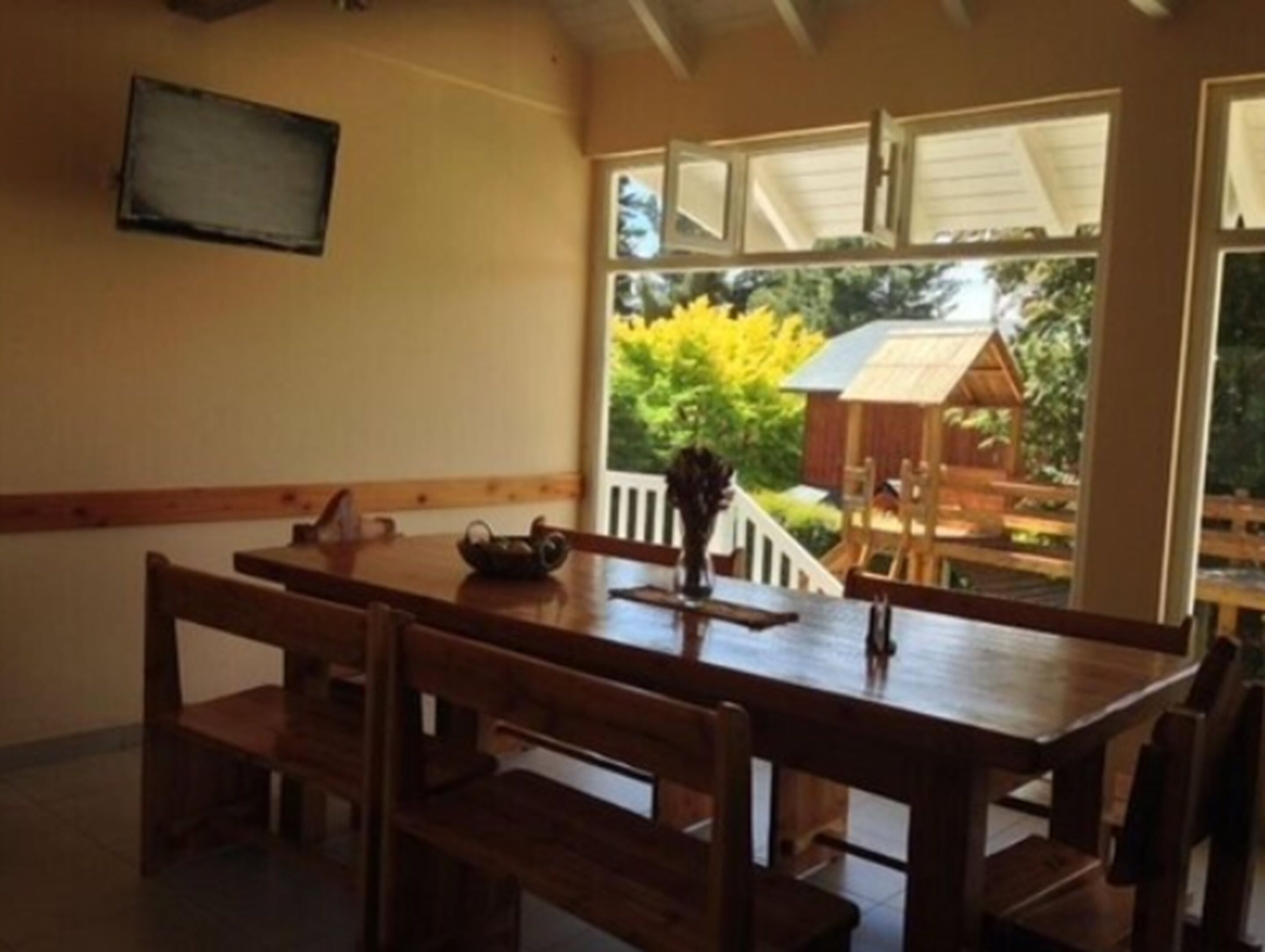A dining area with a wooden table and benches, a television mounted on the wall, and a view of a garden with a wooden structure.