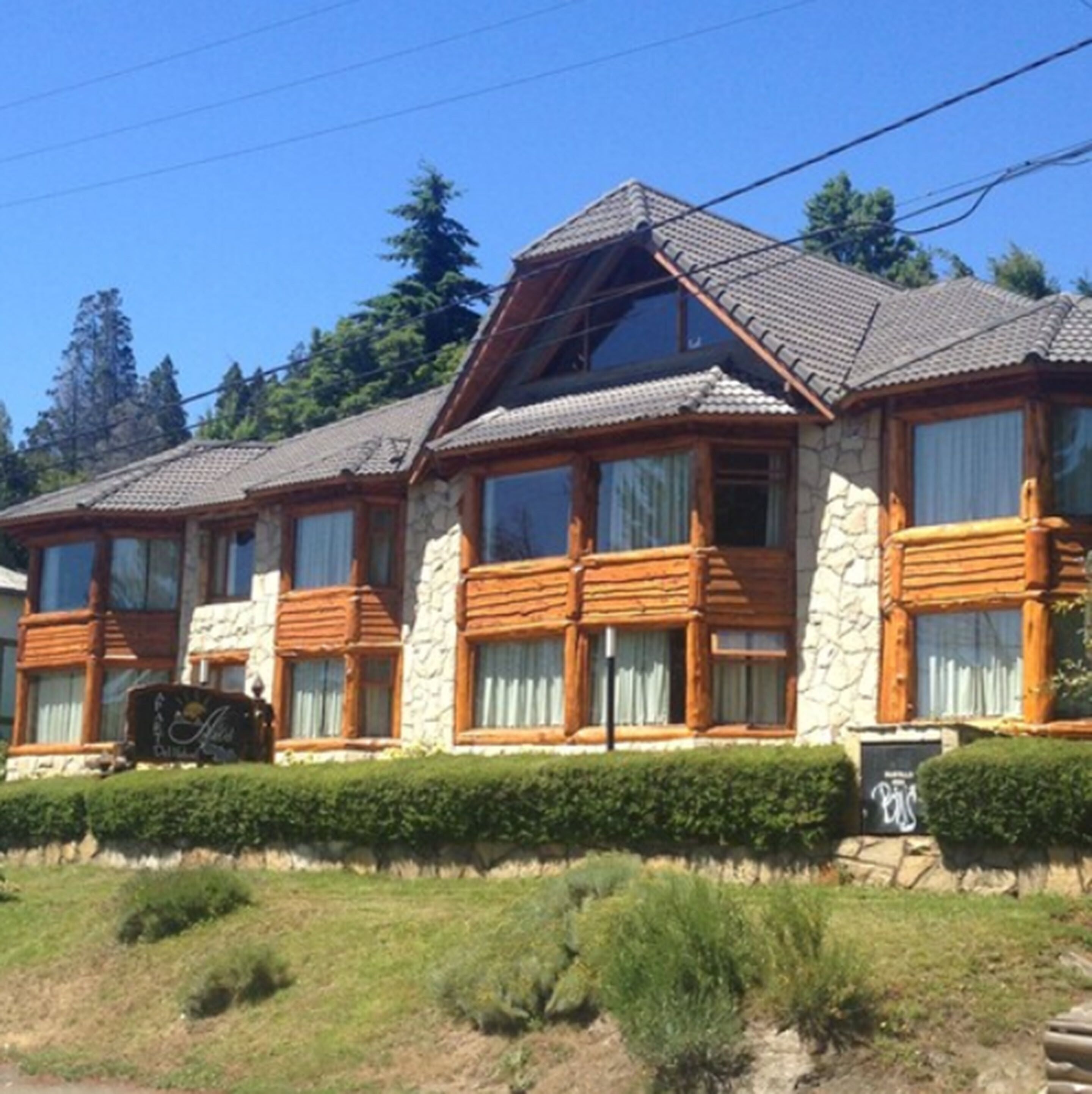 A two-story log cabin with stone walls and wooden windows, surrounded by a well-maintained hedge and a clear sky.