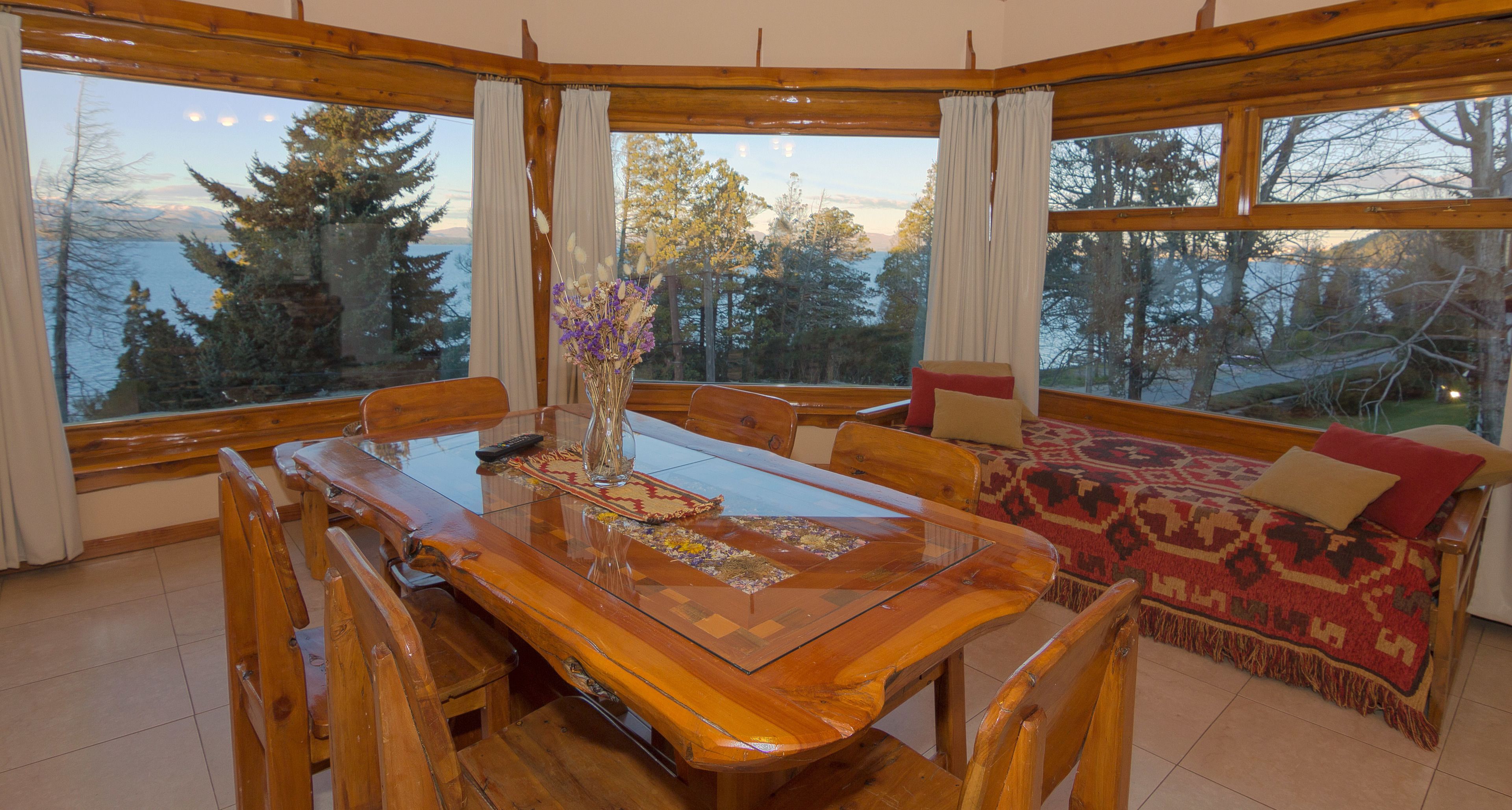 A wooden dining table with chairs, a vase with flowers, and a view of trees and mountains through large windows.