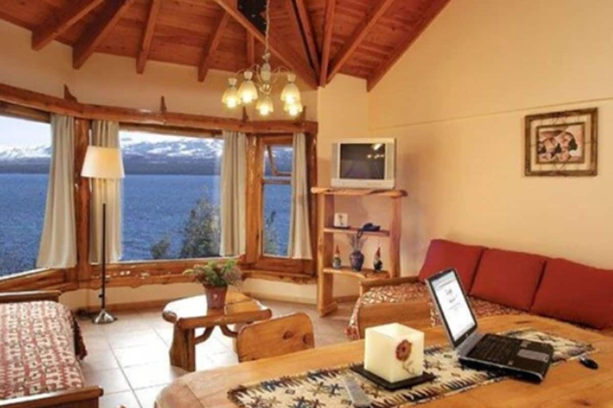 Une pièce chaleureuse avec un plafond en bois, une grande fenêtre donnant sur un lac, un ordinateur portable posé sur une table et un téléviseur sur une étagère.