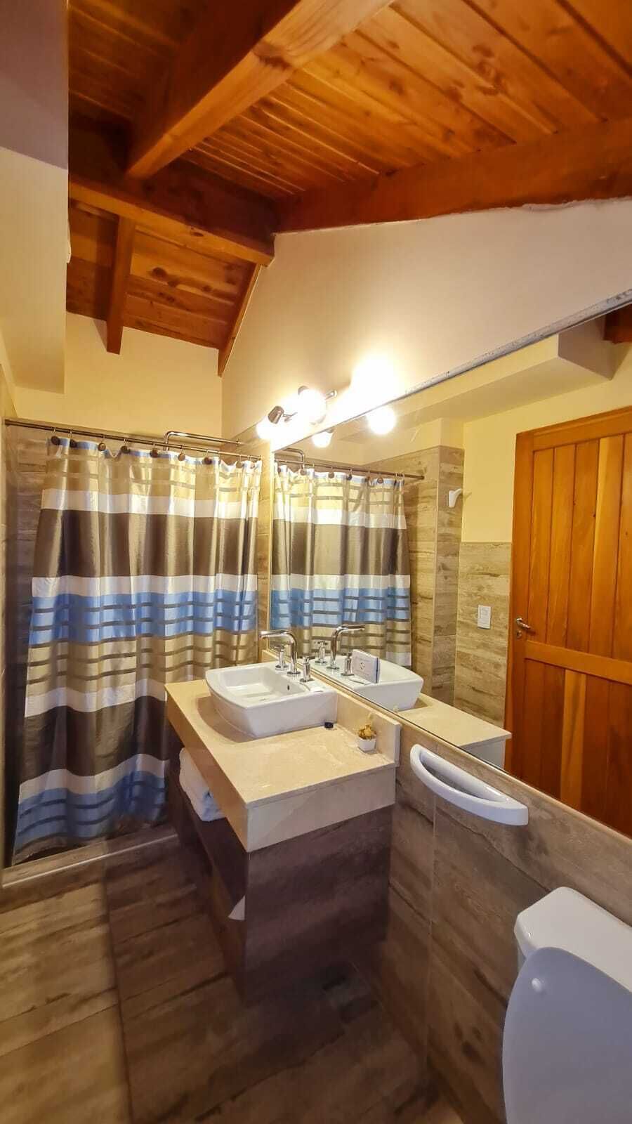 Et badeværelse med træbjælker, en bruseforhæng, to vaske, et spejl og et toilet.