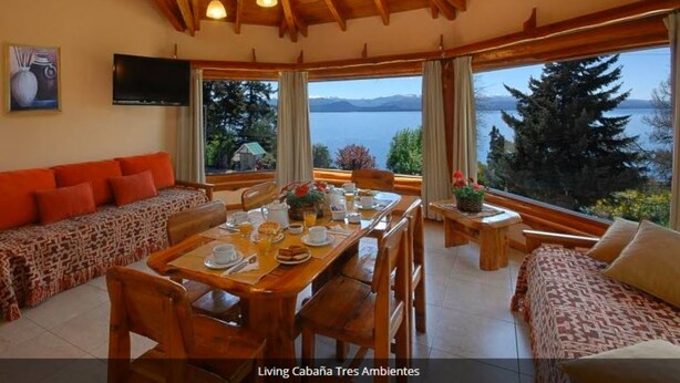 Un comedor con una mesa de madera preparada para cuatro personas, un sofá con cojines estampados y grandes ventanales que ofrecen vistas a un lago y montañas.