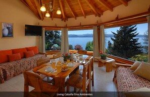 Un comedor con una mesa de madera preparada para cuatro personas, un sofá con cojines estampados y grandes ventanales que ofrecen vistas a un lago y montañas.