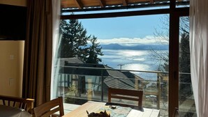 Una mesa de comedor de madera con sillas, un control remoto y un cuenco de fruta sobre un mantel con estampado. Un ventanal con cortinas ofrece vistas a un paisaje montañoso.