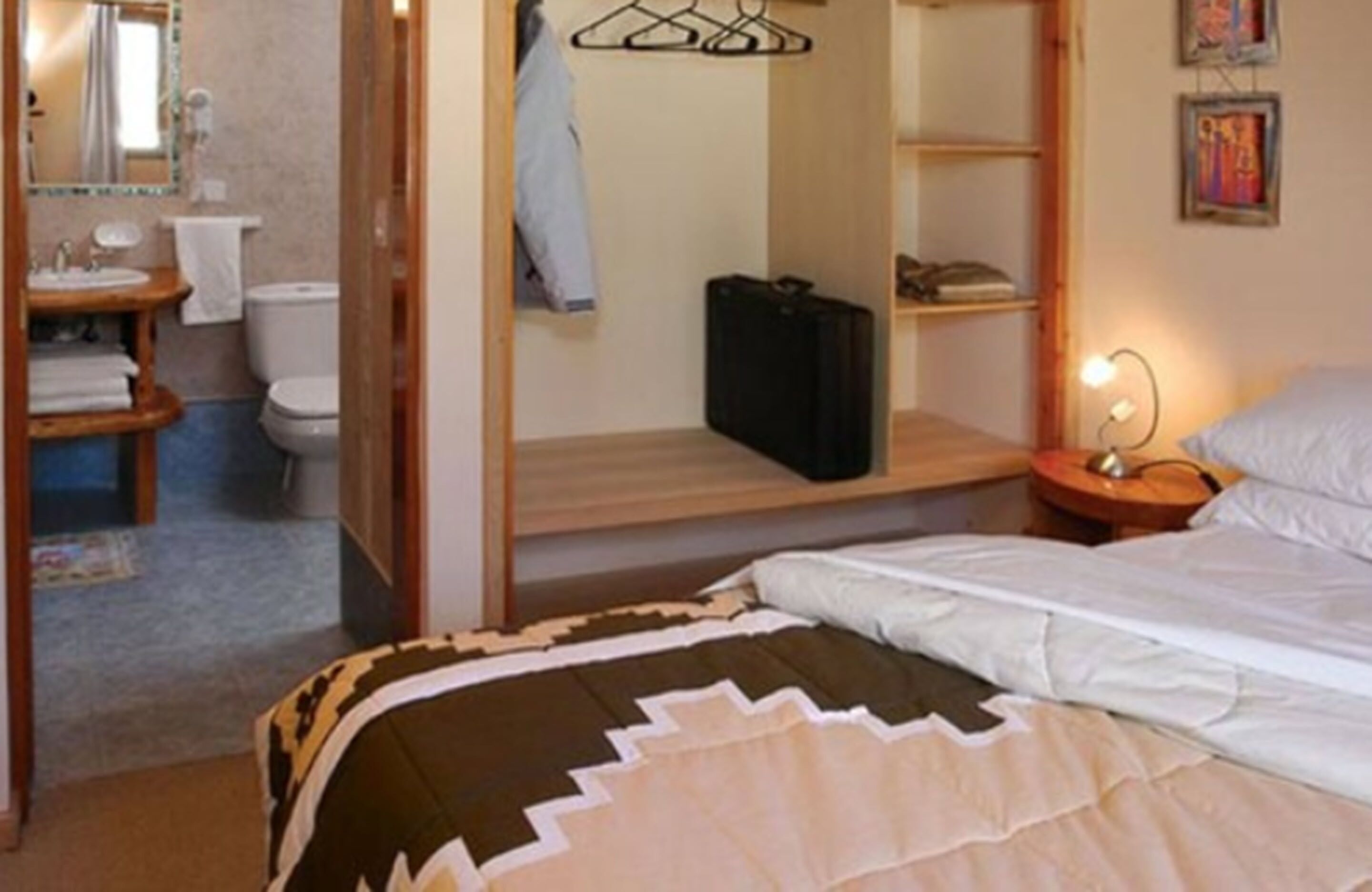 Un dormitorio con cama, mesita de noche con lámpara, perchero, armario y baño visible a través de una puerta abierta.