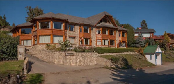 A large, multi-story wooden house with stone foundations, a stone staircase leading to the entrance, and a small adjacent structure.