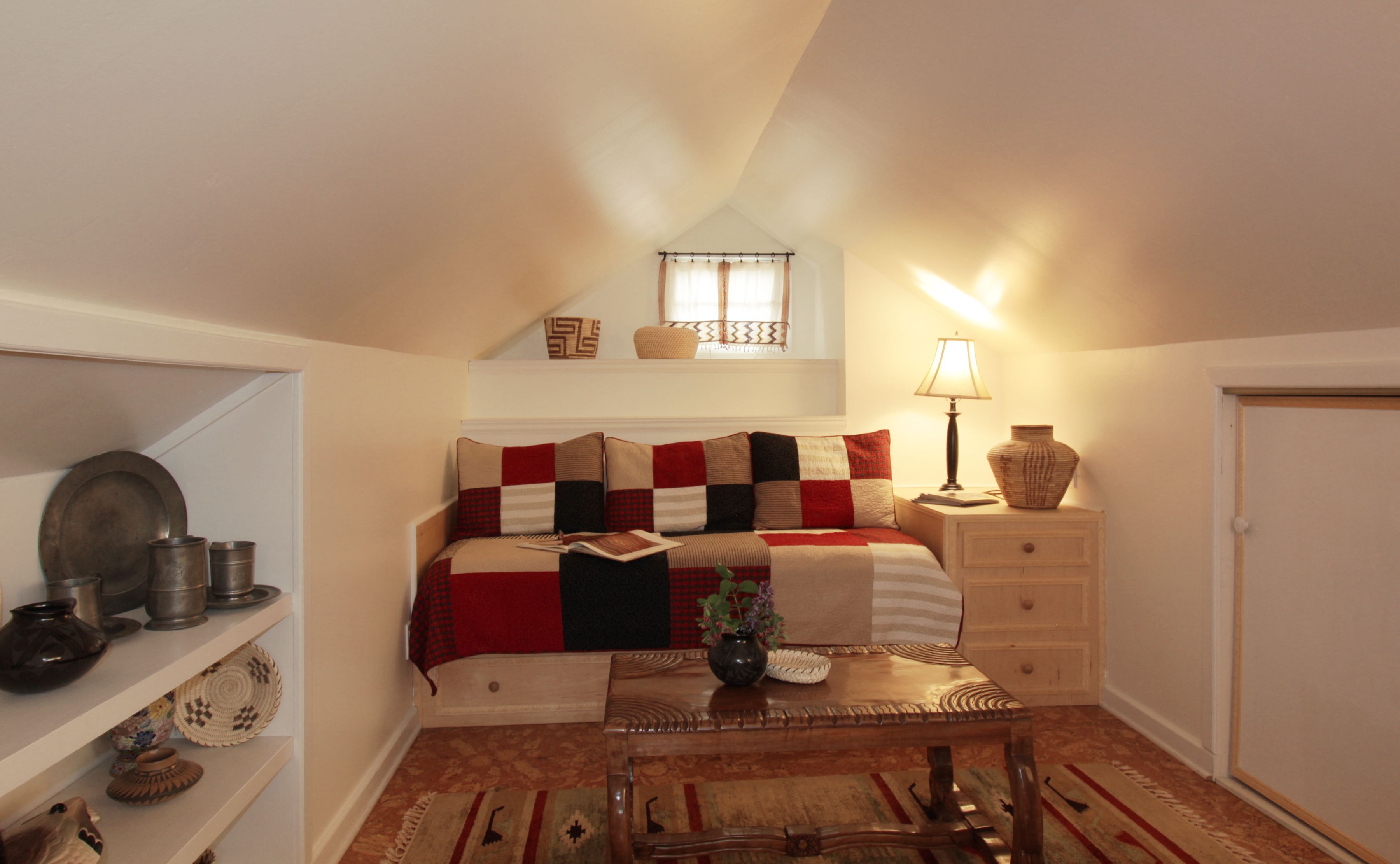 A cozy loft bedroom with a sloped ceiling, a bed with a red and black checkered blanket, a bedside table with a lamp, and a small window with a curtain.