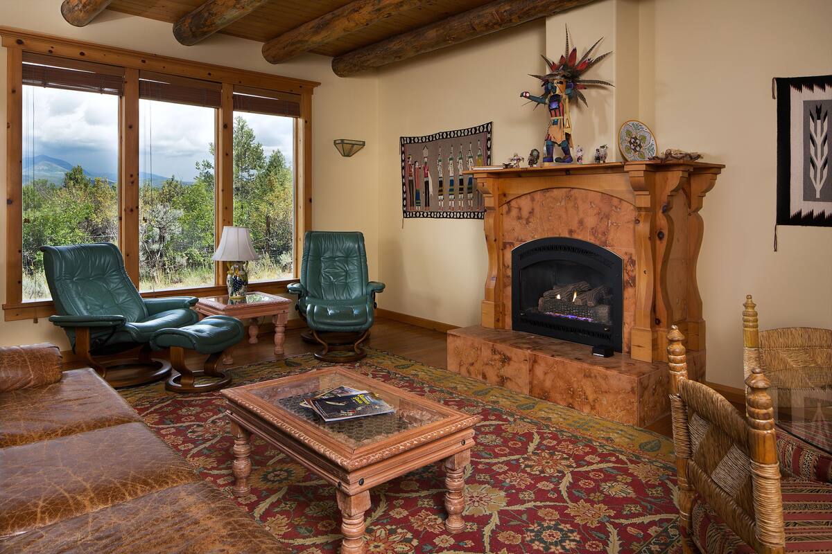 Un salon chaleureux avec une cheminée, des fauteuils en cuir, une table basse et une grande fenêtre offrant une vue panoramique.