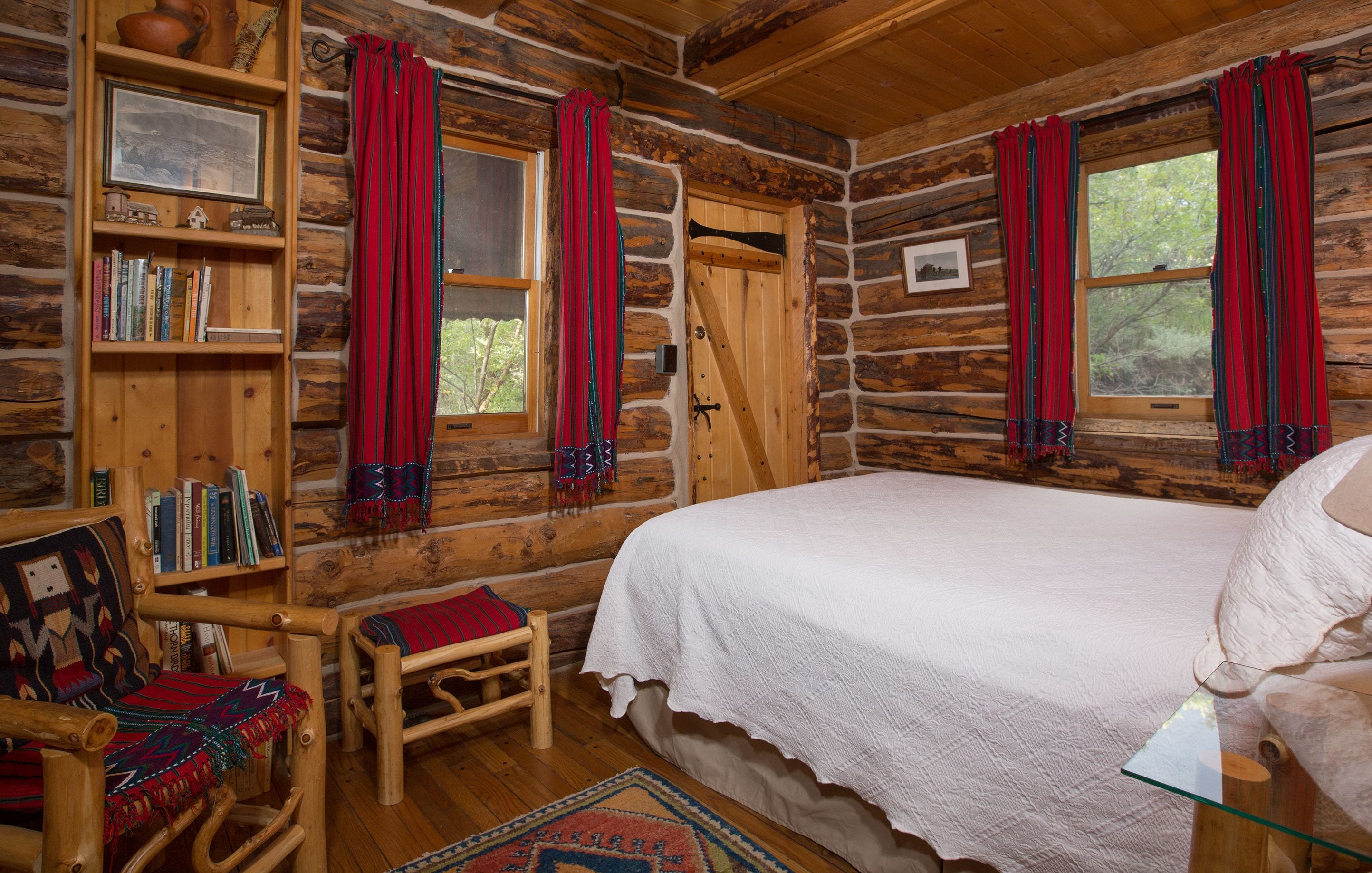 Une chambre aménagée dans un chalet en rondins, avec un lit, des étagères en bois remplies de livres, un fauteuil à bascule et une fenêtre ornée de rideaux rouges.