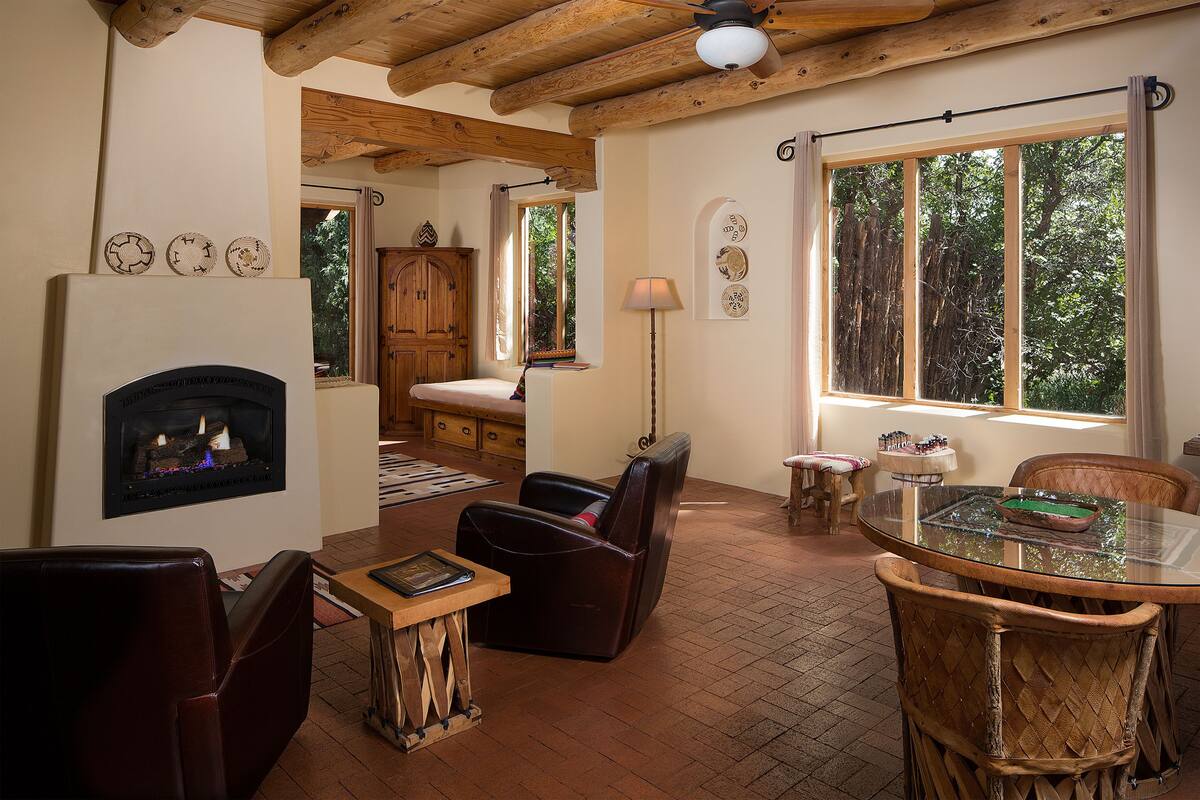 Un salon chaleureux avec une cheminée, des fauteuils en cuir, une table à manger et de grandes fenêtres donnant sur l’extérieur.
