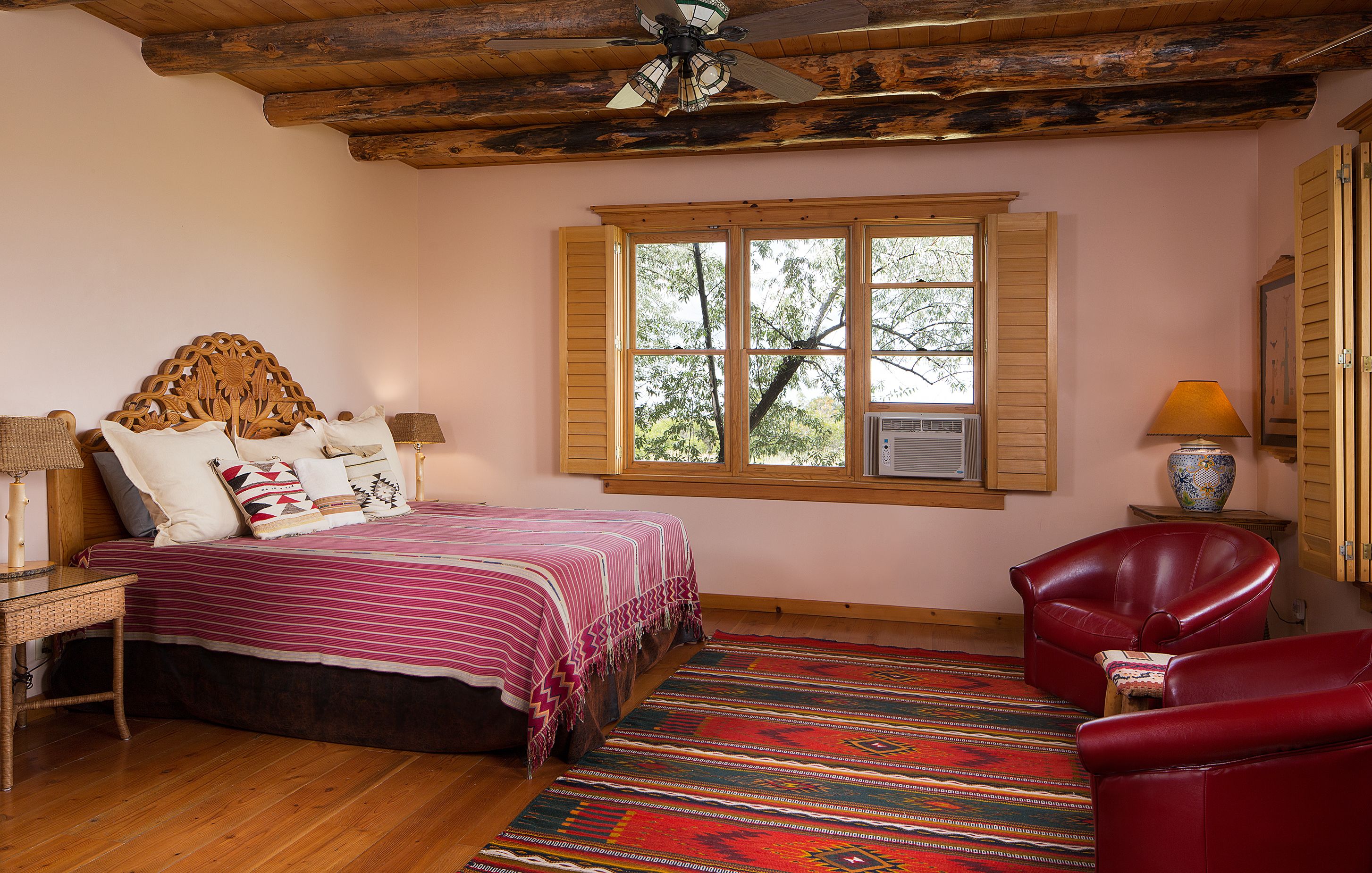 A bedroom with a wooden ceiling, a bed with a patterned bedspread, a red leather chair, and a window with shutters.
