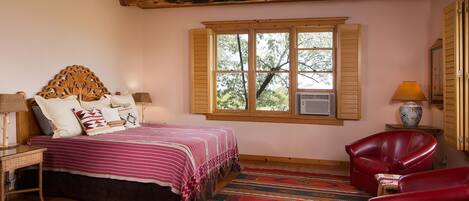 A bedroom with a wooden ceiling, a bed with a patterned bedspread, a red leather chair, and a window with shutters.