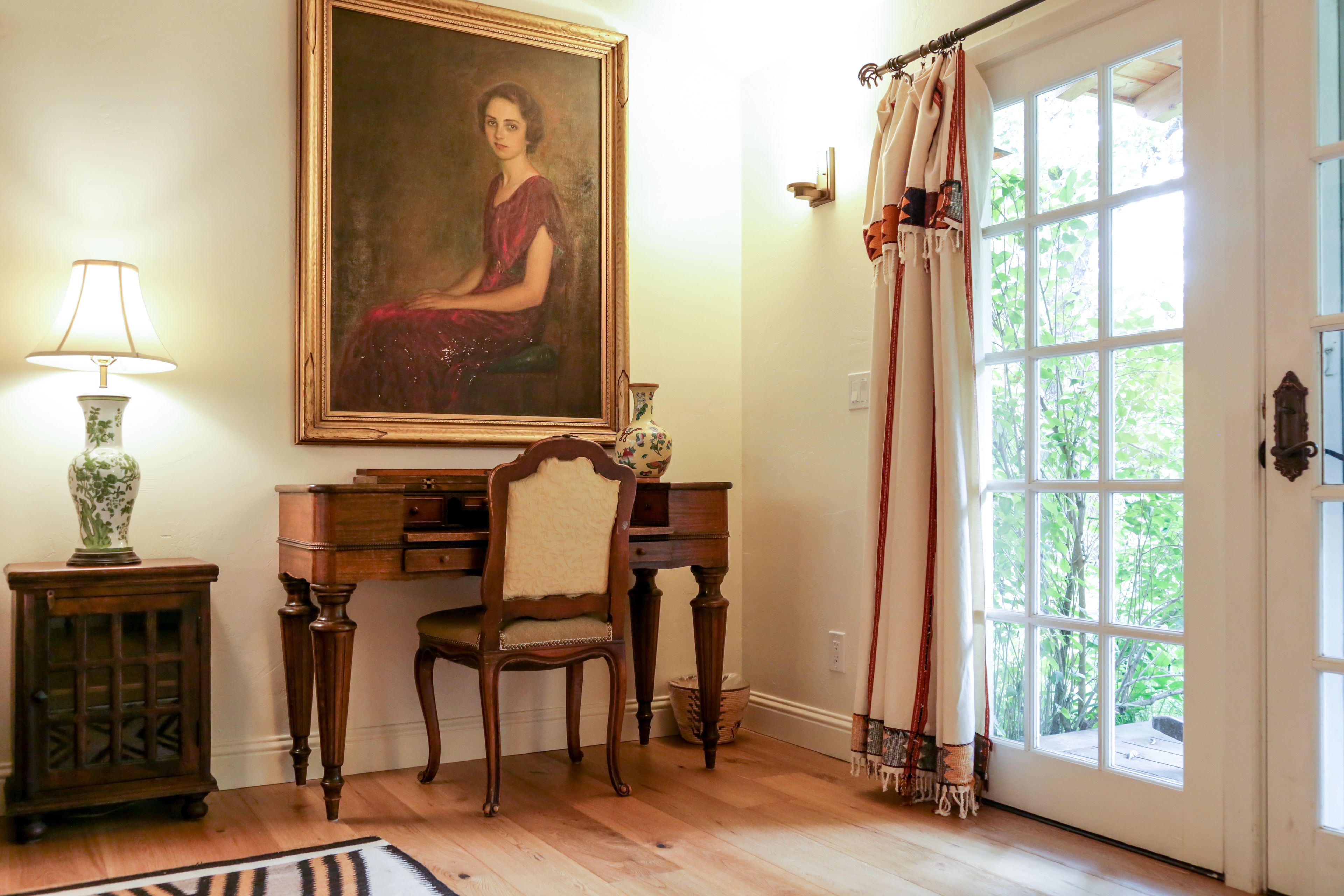 A room with a wooden desk, a chair, a lamp, a painting, and a window with curtains.