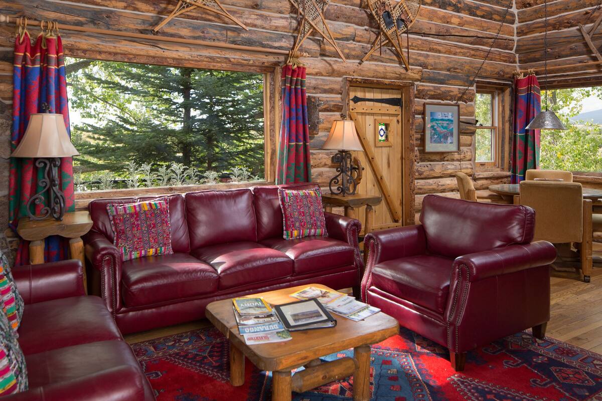 L’intérieur d’une cabane en rondins est aménagé avec un canapé en cuir rouge, une table basse en bois sur laquelle sont posés des magazines, et offre une vue sur les arbres à travers de grandes fenêtres.