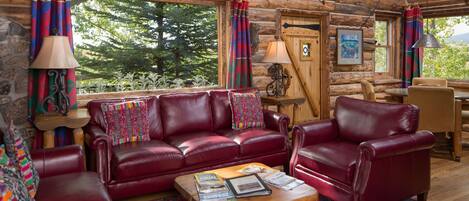 A log cabin interior with a red leather sofa set, a wooden coffee table with magazines, and a view of trees through large windows.