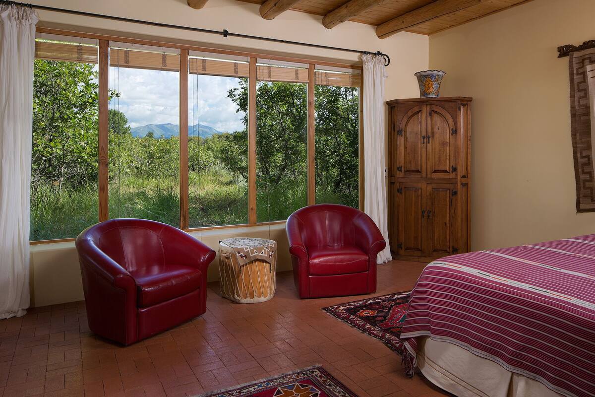Une chambre avec une grande fenêtre, un lit, deux chaises rouges, une armoire en bois et un tapis à motifs.