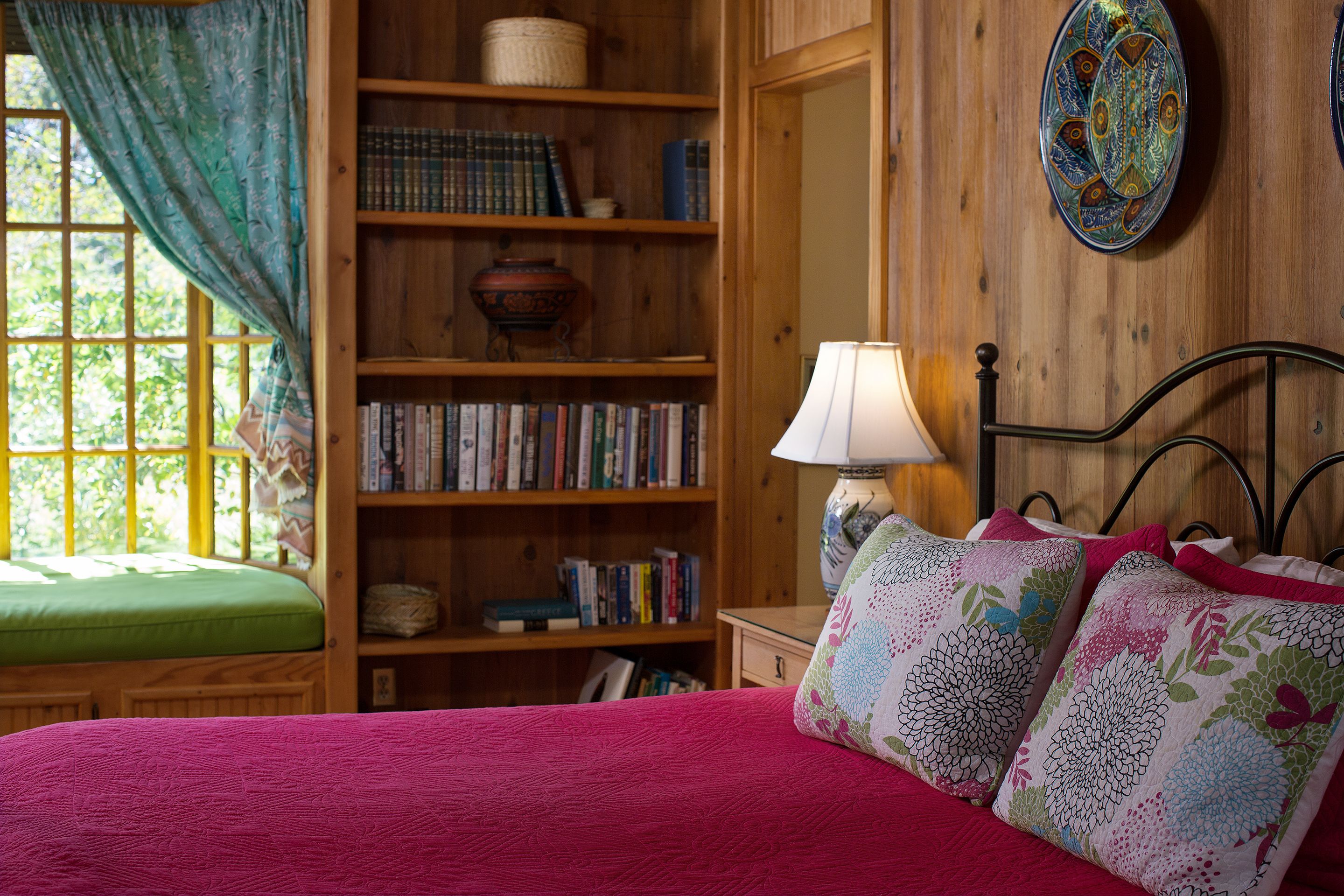 A cozy room with a wooden bookshelf, a green cushioned bench, and a bed with a red blanket and floral pillows.