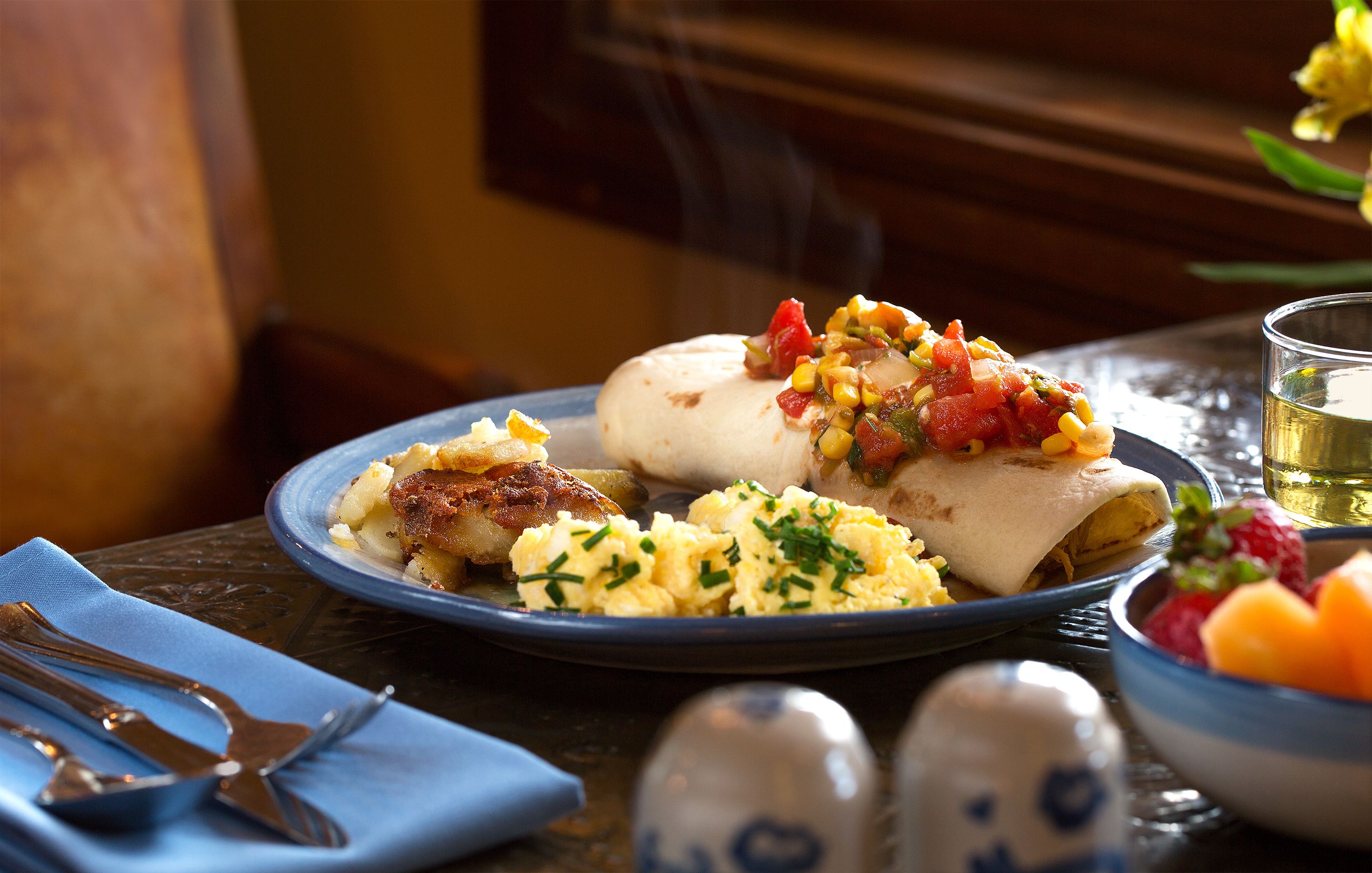 Un plato con alimentos, incluyendo un rollo, huevos revueltos y un acompañamiento, sobre una mesa con un mantel azul y saleros y pimenteros.