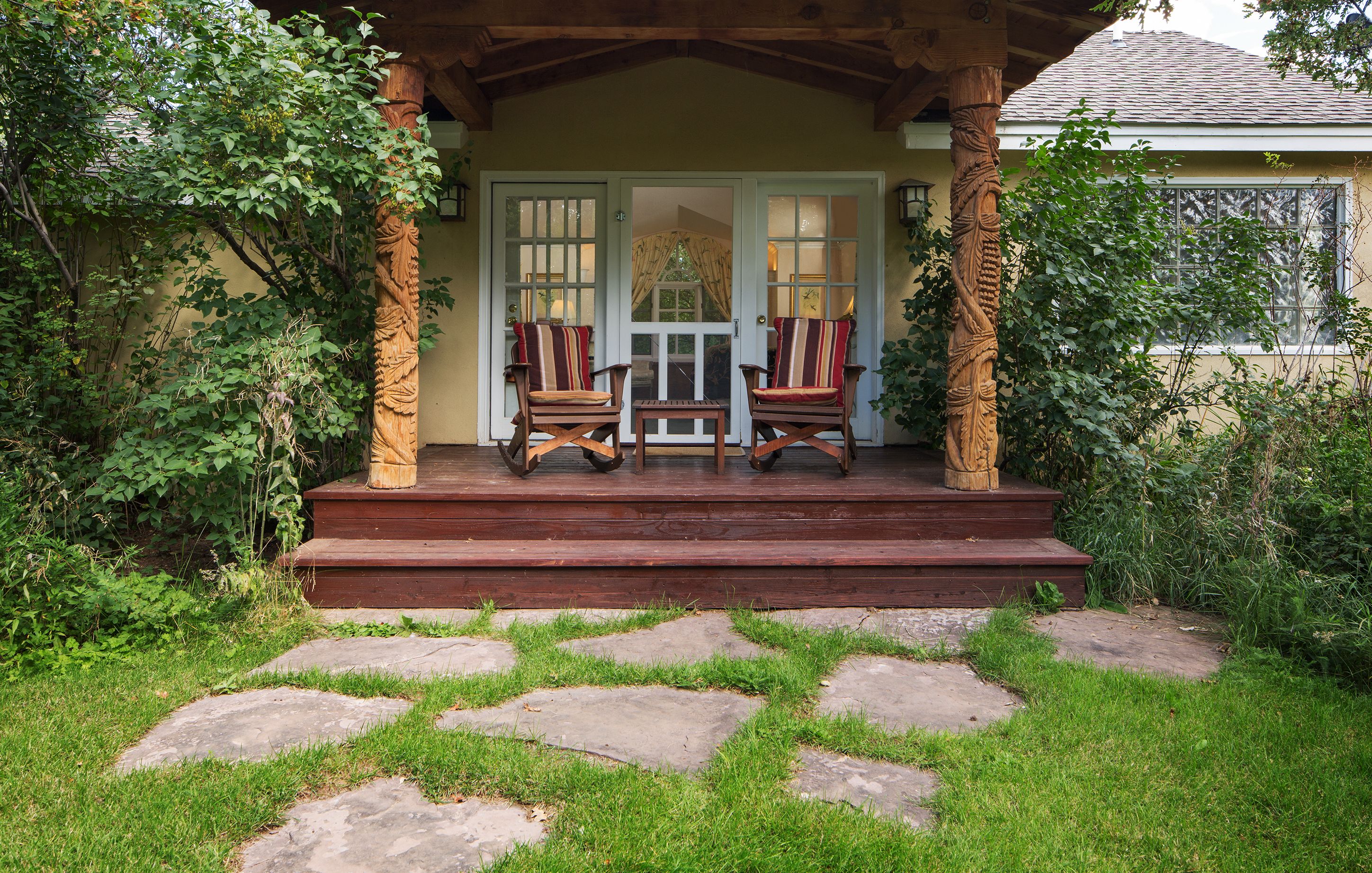 Een houten veranda met twee stoelen en een tafel, omgeven door begroeiing en stenen trappen.