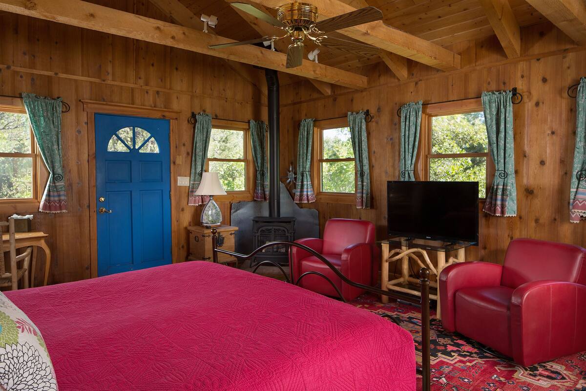 Une pièce de style cabane en bois, avec un grand lit, une télévision, deux fauteuils rouges, une cheminée et un ventilateur de plafond.