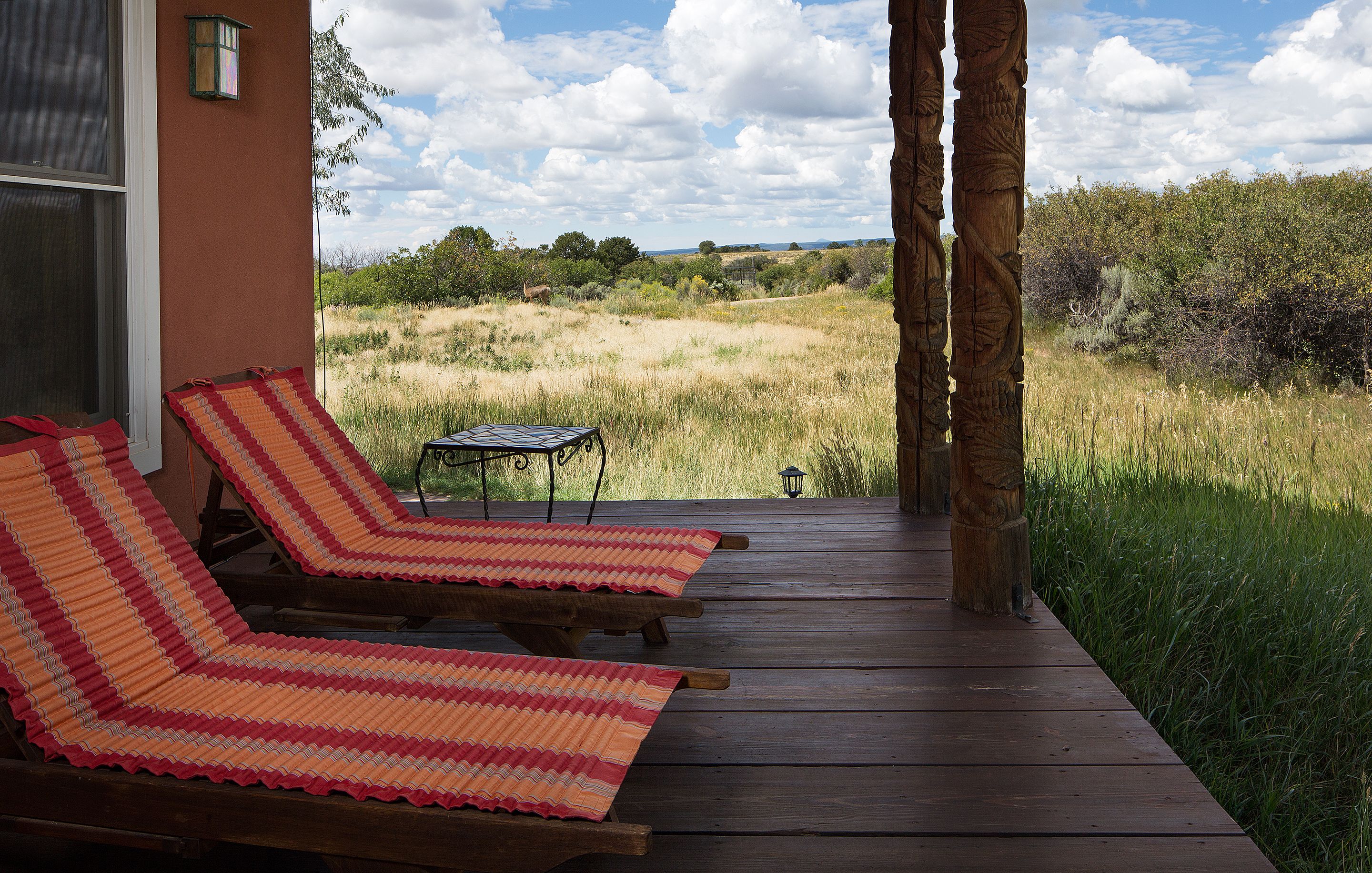 Terraza de madera con dos sillones de descanso y una mesa, con vistas a un campo cubierto de hierba y árboles.