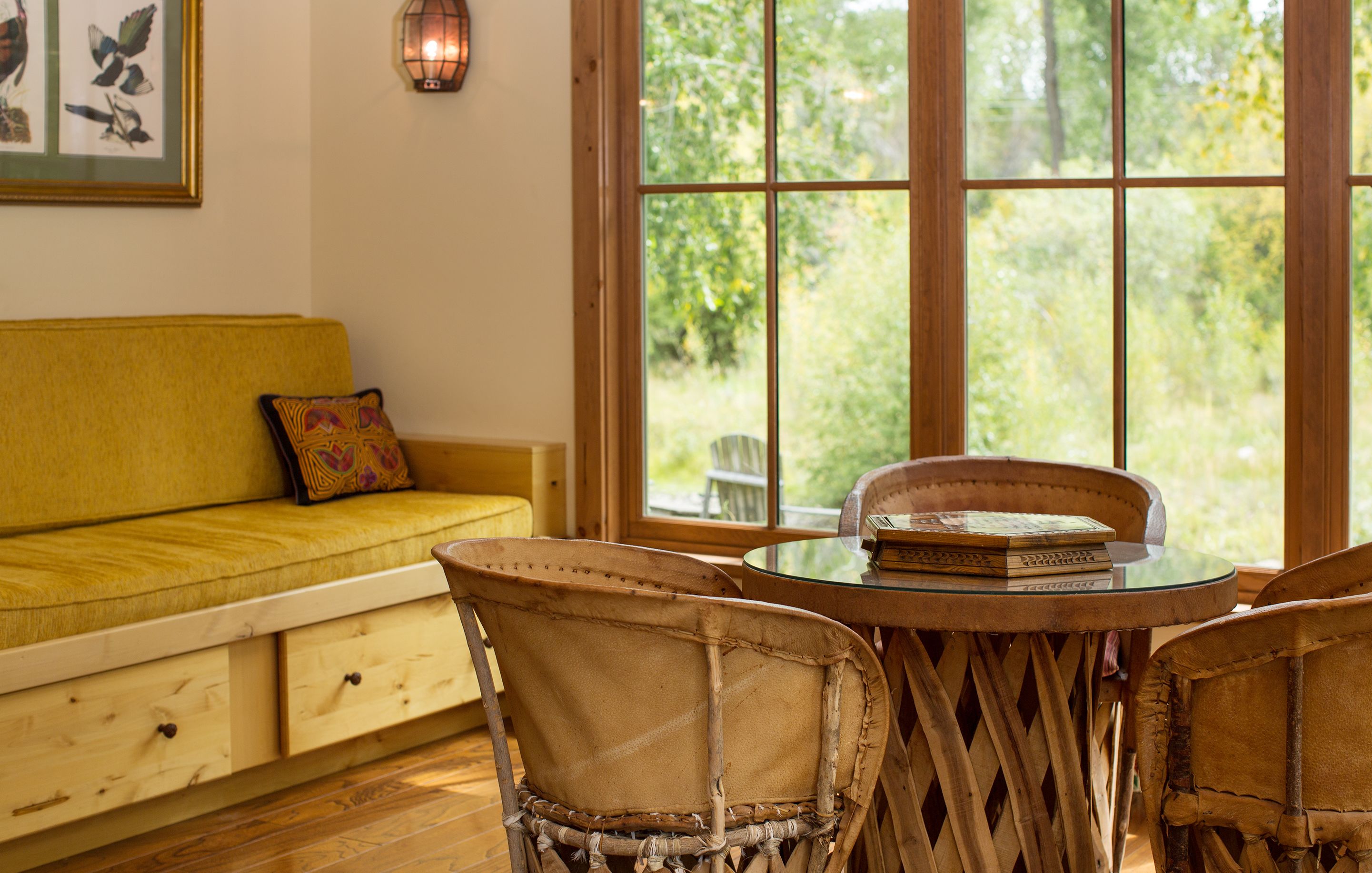 Ein Zimmer mit Holzboden, einem gelben Sofa mit Kissen, Korbstühlen, einem Glastisch und einem Fenster mit Blick auf Grünflächen.