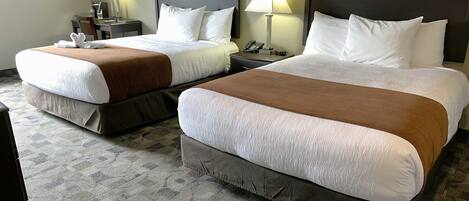 A hotel room with two beds, each with a brown headboard and white bedding. There are two bedside tables with lamps, a framed picture on the wall, and a carpeted floor.