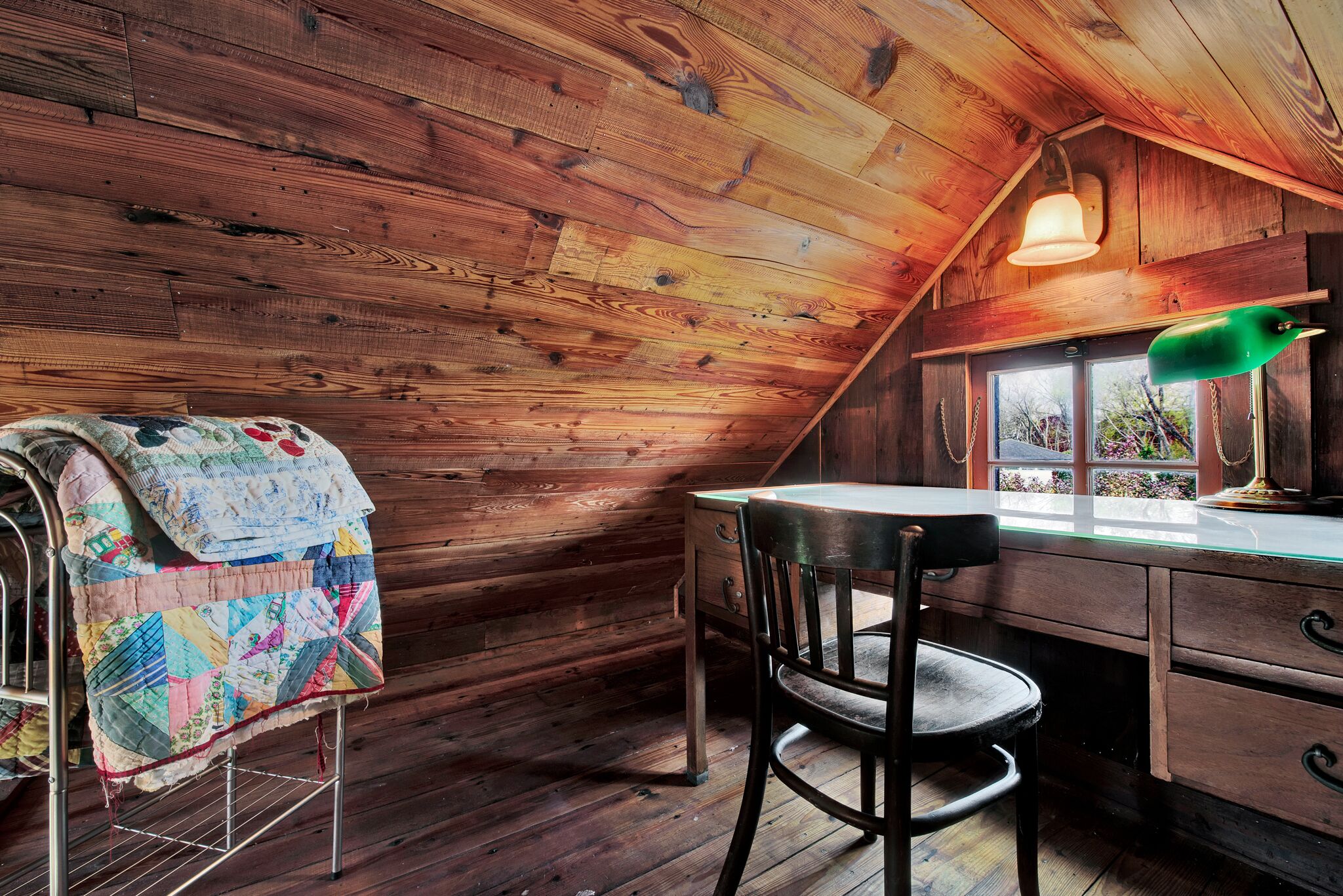 Una habitación cálida tipo cabaña con interior de madera, un escritorio con una silla, un edredón acolchado sobre una silla y una ventana con vistas.