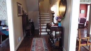 Un couloir avec un plancher en bois, un tapis à motifs, un escalier, une table ornée d’un vase de fleurs et une chaise.